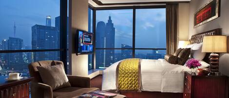 A hotel room with a large window offering a city view, a bed with a yellow accent pillow, a brown armchair, a bedside table with a lamp, and a flat-screen TV mounted on the wall.