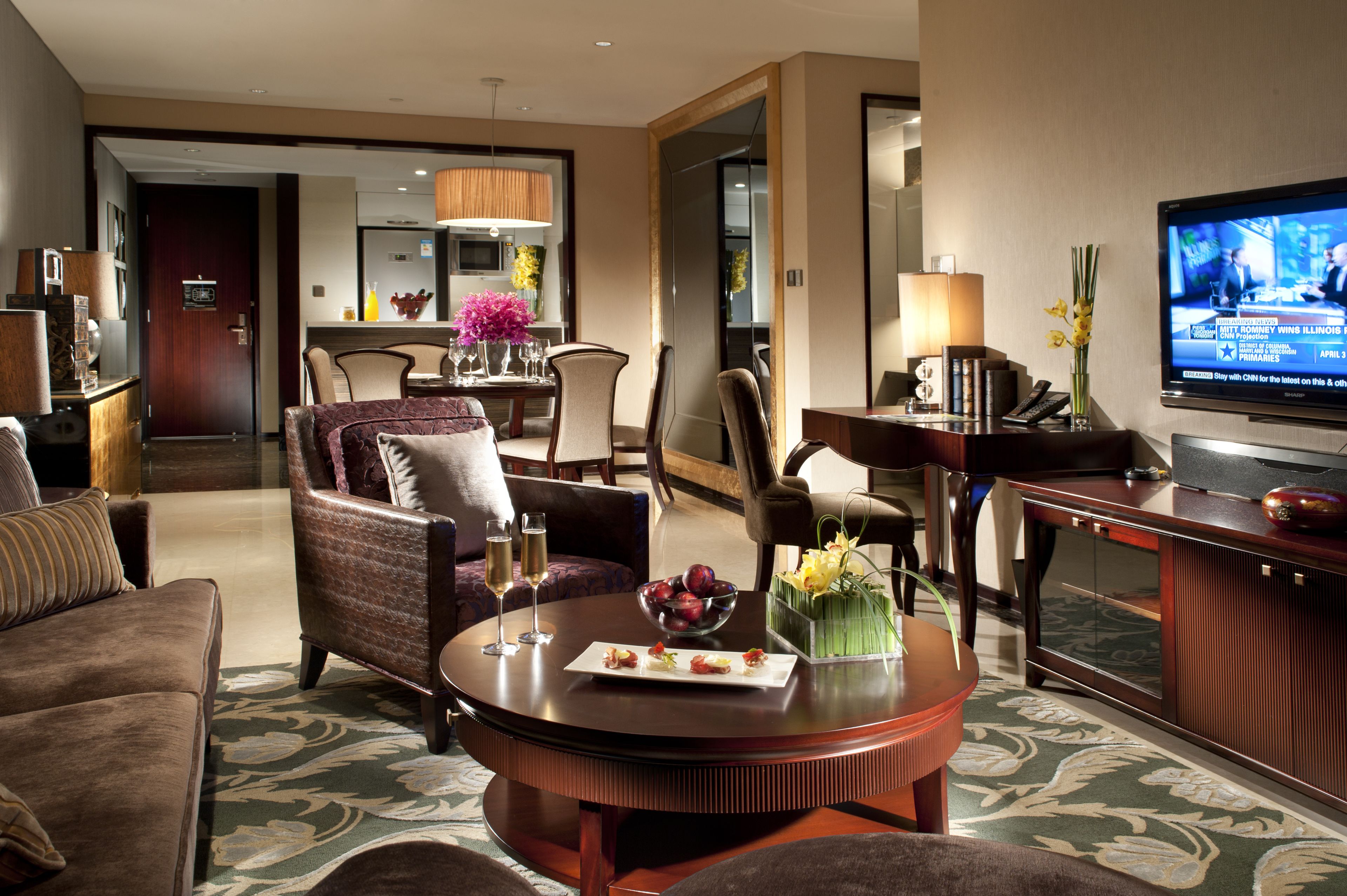 Une chambre d’hôtel dotée d’un espace salon comprenant un canapé, des fauteuils, une table basse avec des collations et des boissons, une télévision, ainsi qu’un coin repas avec une table et des chaises.