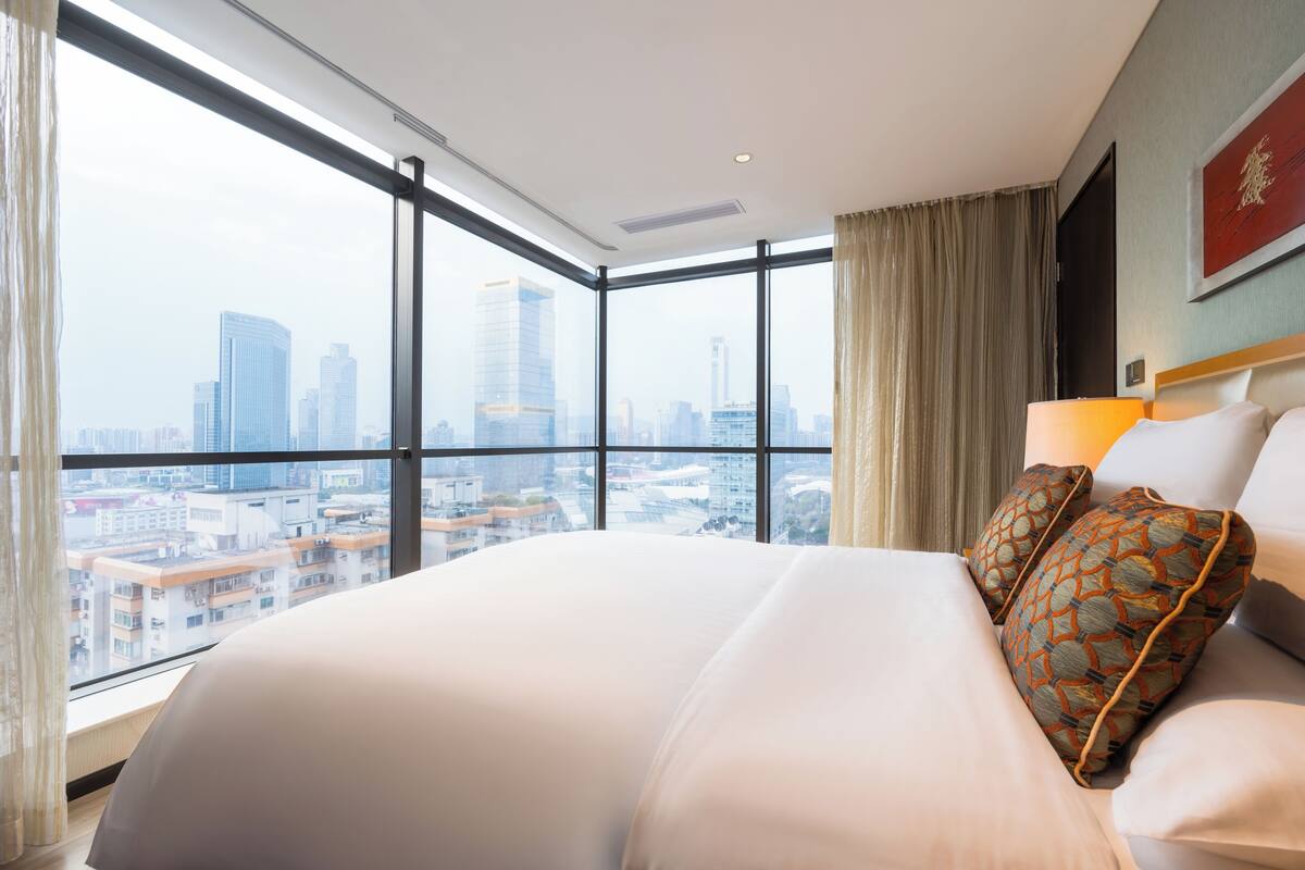 A hotel room with a large bed, a view of a cityscape through floor-to-ceiling windows, and a city skyline in the background.