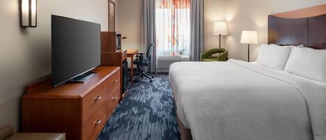 A hotel room with a bed, a television on a wooden dresser, a desk with a chair, and a window with curtains.