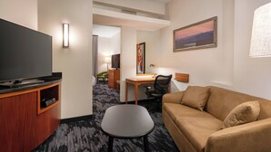 A hotel room with a flat-screen TV, a wooden desk, a black chair, a brown sofa, and a painting on the wall. - Fairfield Inn & Suites Visalia Tulare (Tulare)