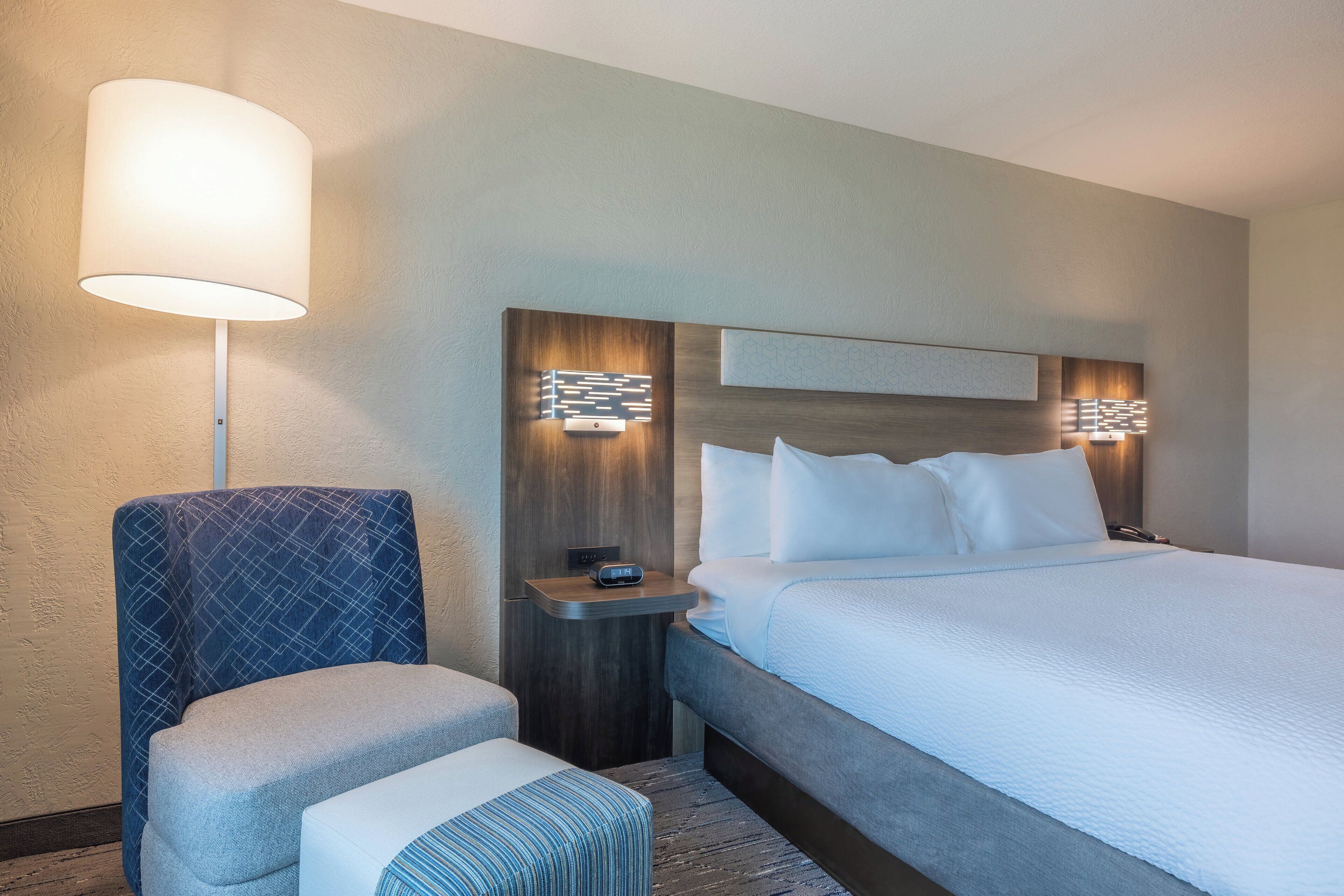 A hotel room with a large bed, a bedside table with a lamp, a chair, and a small ottoman.