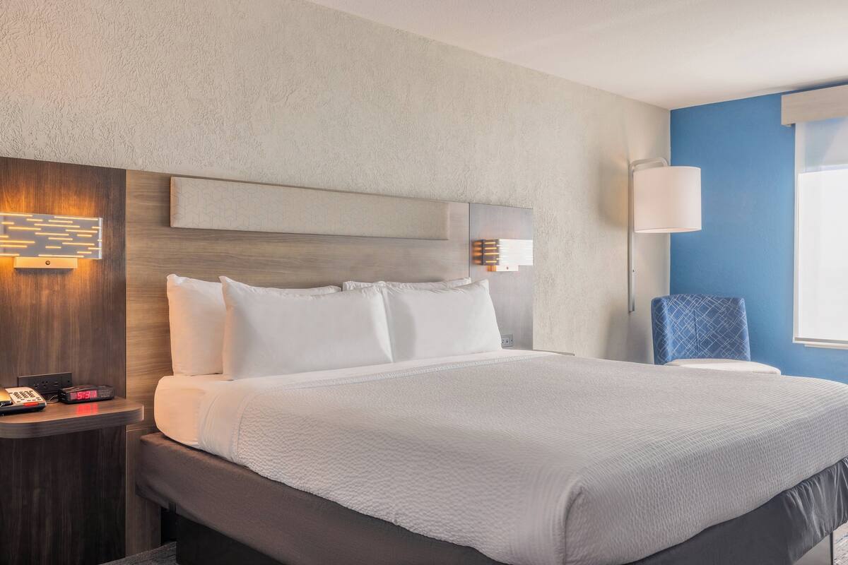 Una habitación de hotel con una cama grande, una mesita de noche con teléfono y reloj, una lámpara de pared y una silla.