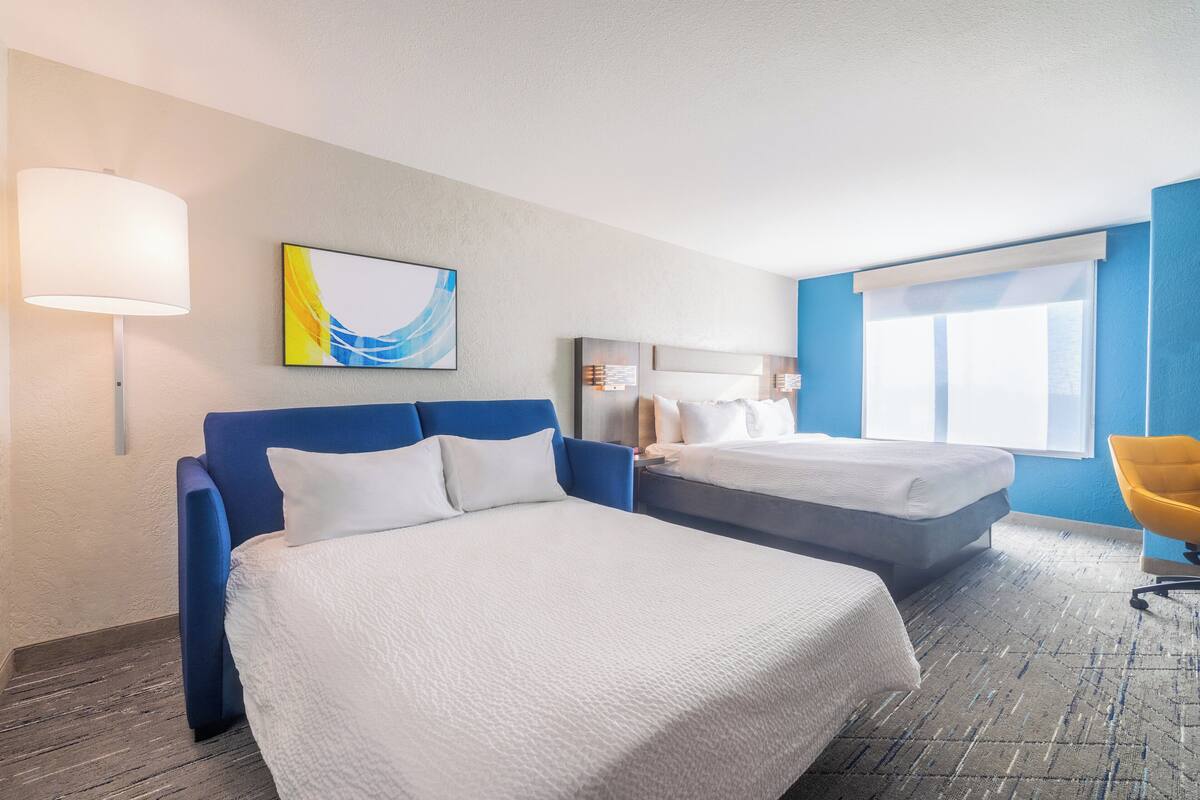 Habitación de hotel con dos camas, un escritorio, una silla, una lámpara y un cuadro en la pared.
