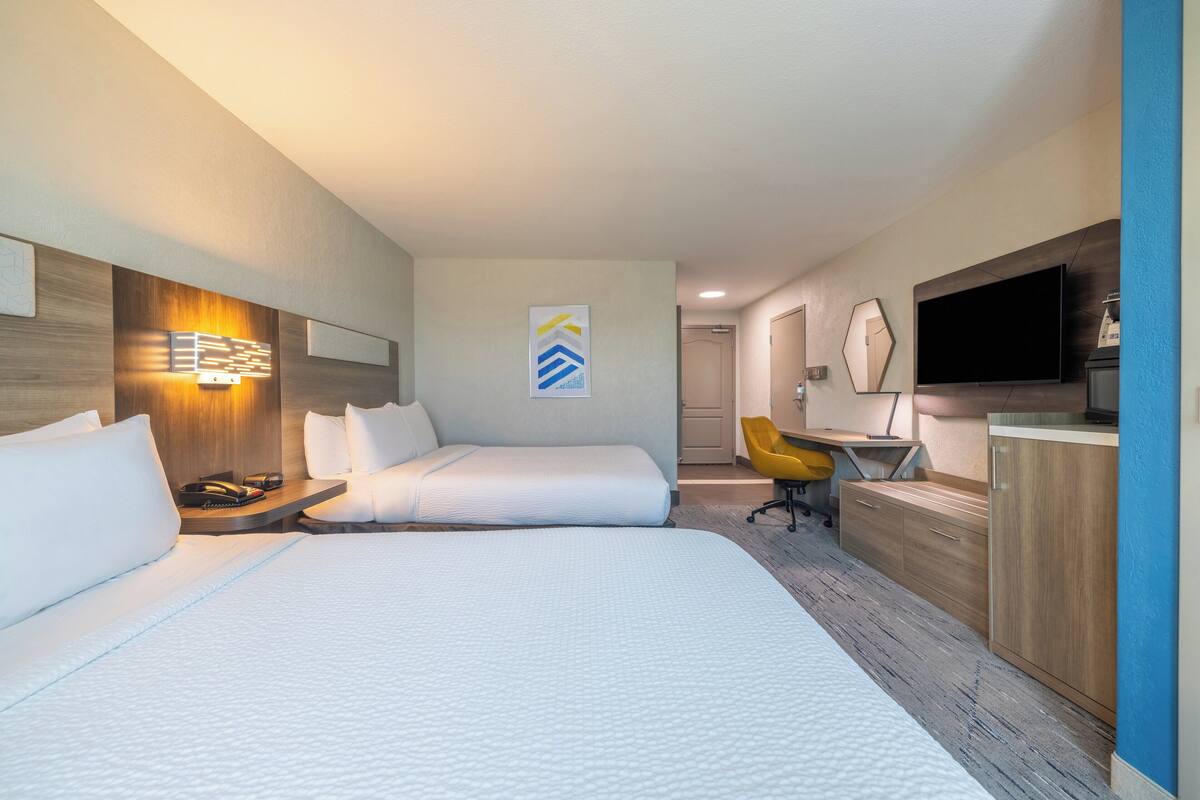 Habitación de hotel con dos camas, un escritorio, una silla, un televisor y un baño al fondo.
