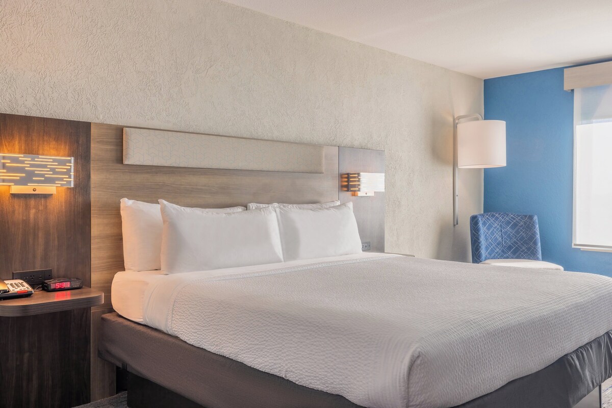 Una habitación de hotel con una cama grande, una mesita de noche con teléfono y reloj, una lámpara de pared y una silla.