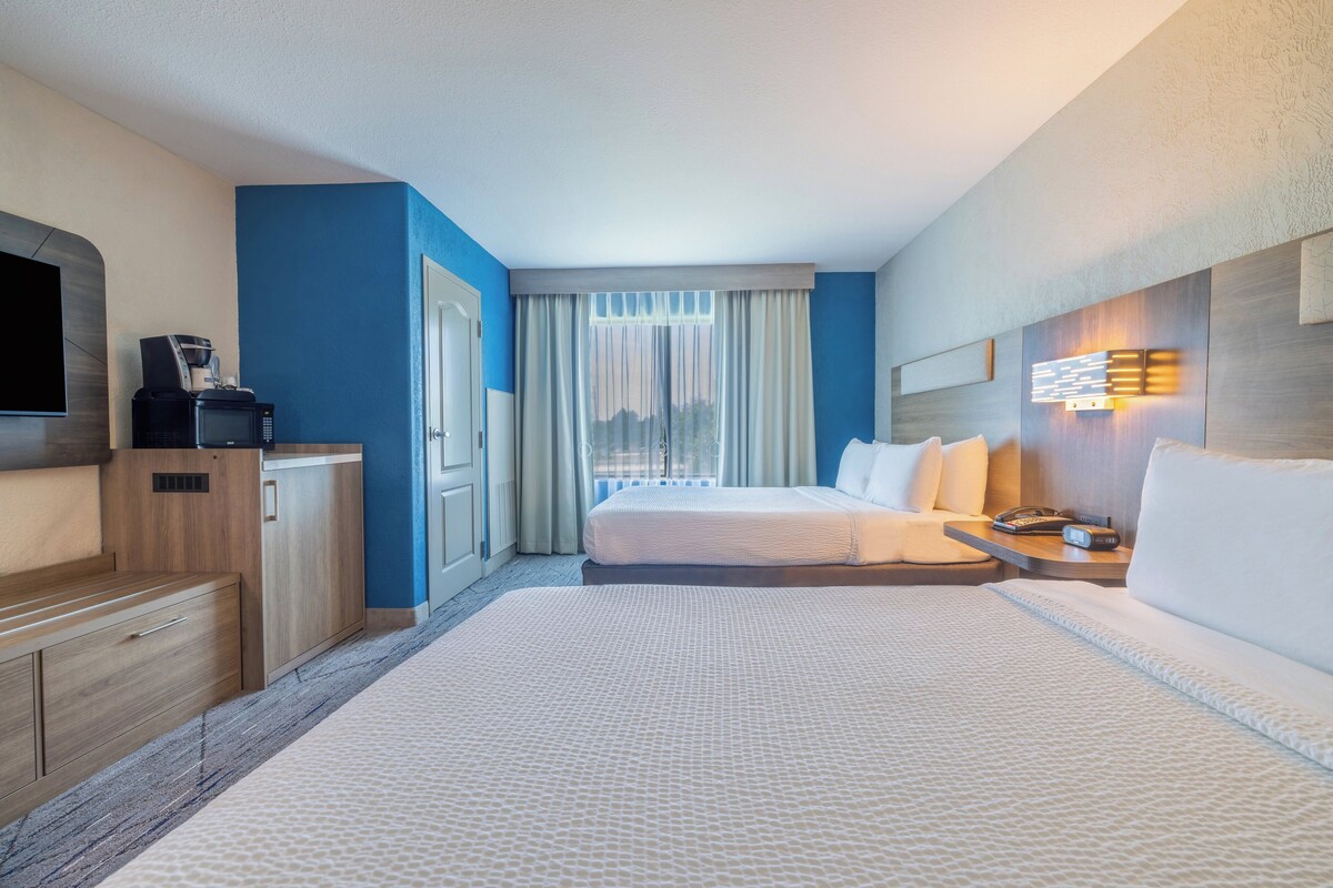 Una habitación de hotel con cama, escritorio, televisor, cafetera y ventana con cortinas.