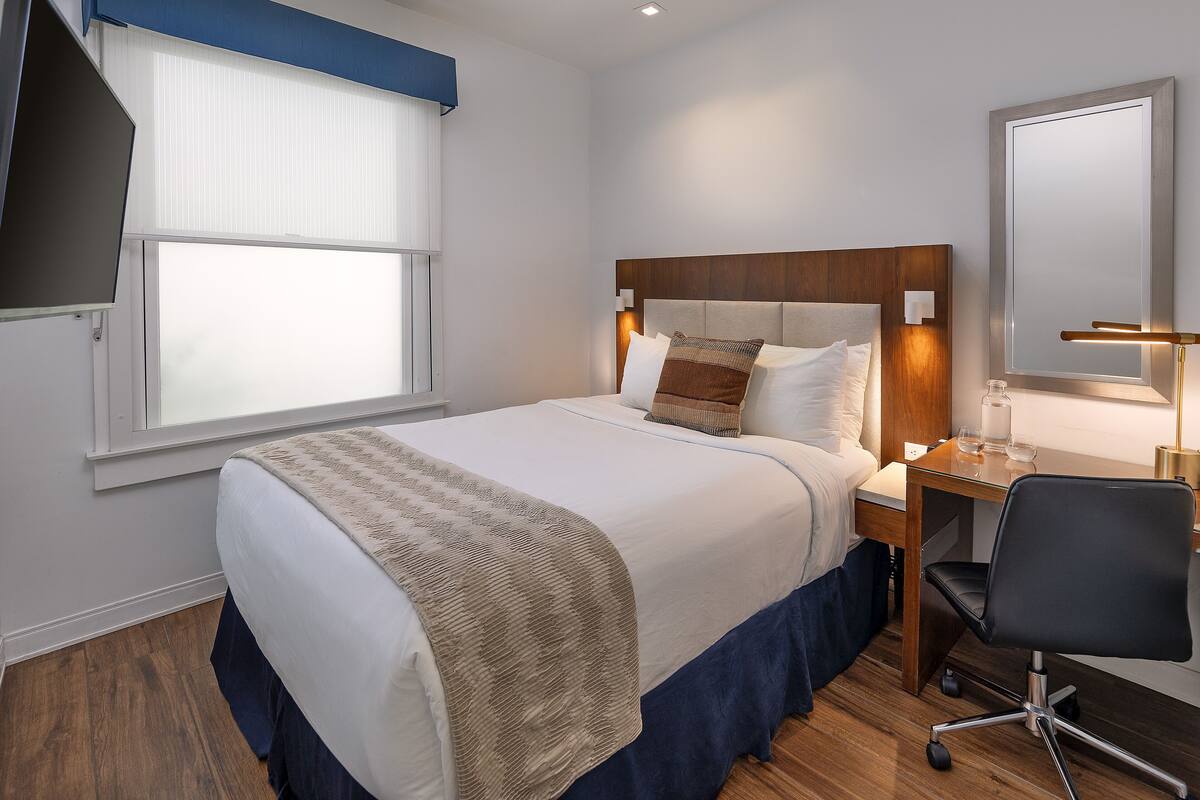 Quarto de hotel bem arrumado com cama, mesa de cabeceira, cadeira e uma janela grande com persianas.