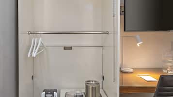 Una kitchenette compacta con cafetera, microondas y tetera, un escritorio pequeño con lámpara, y un televisor montado en la pared.