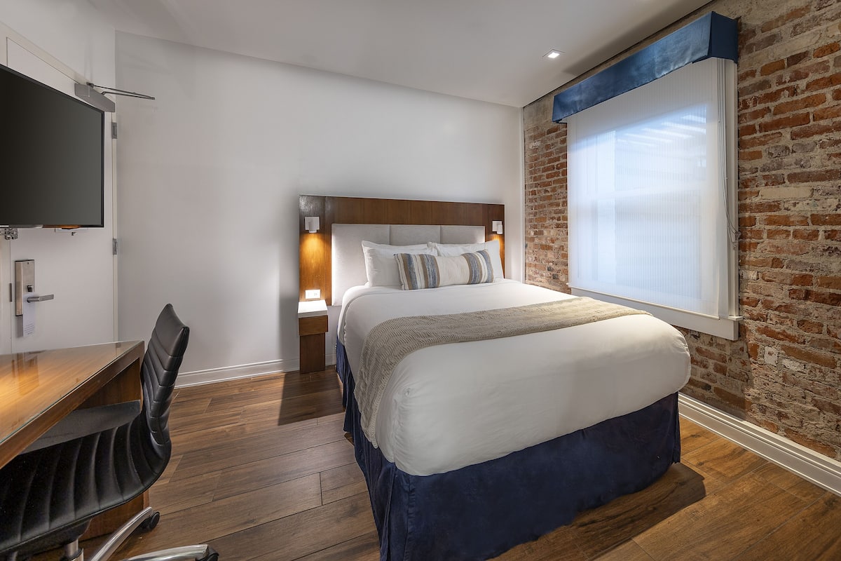 Quarto de hotel com cama, escrivaninha, cadeira, televisão e parede de tijolos.