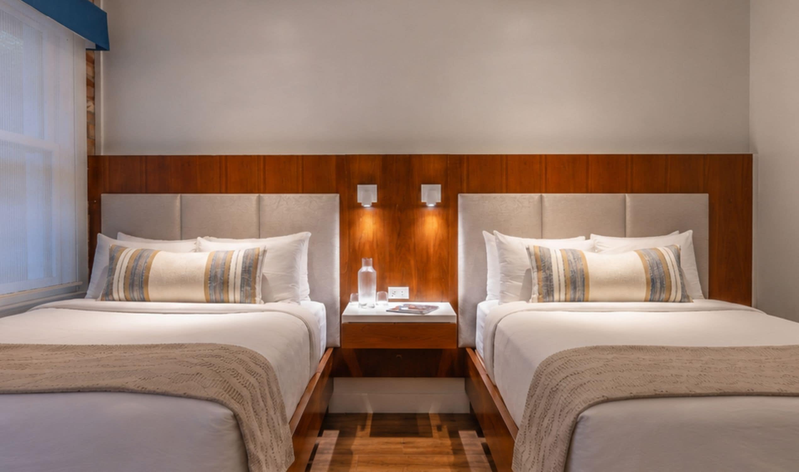A hotel room with two beds, a wooden headboard, a nightstand with a lamp, and a window with blinds.
