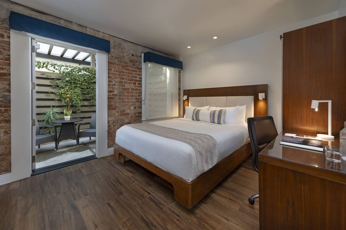 Quarto de hotel com uma cama grande, uma escrivaninha com cadeira, um armário de madeira e uma porta de vidro corrediça que dá para uma área externa com plantas.