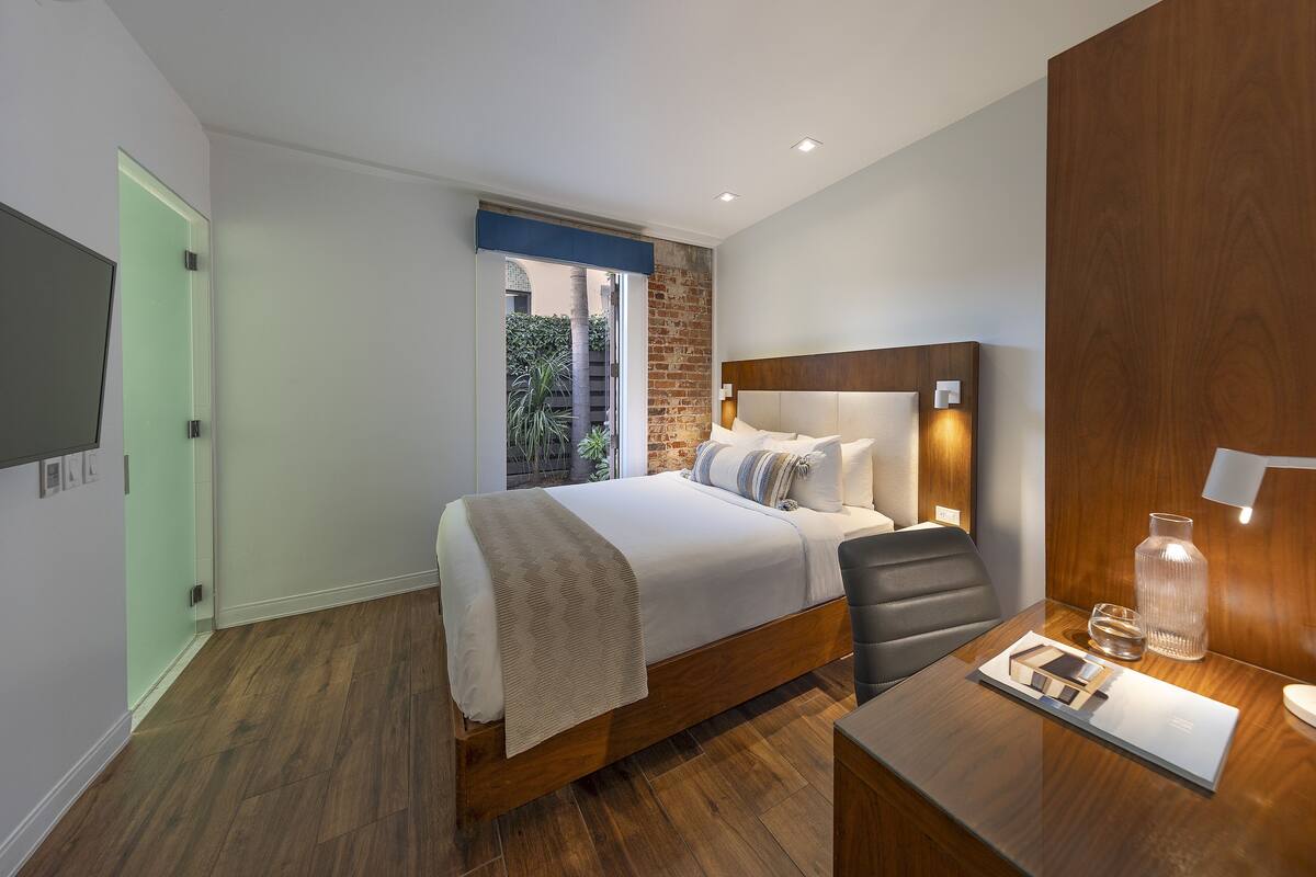 Quarto de hotel moderno com cama de madeira, escrivaninha com abajur e vista para o verde lá fora.