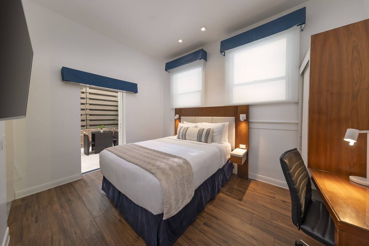 Quarto de hotel moderno com uma cama grande, piso de madeira, uma escrivaninha com cadeira e uma janela com persianas.