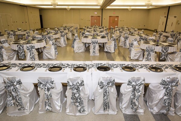 Uma sala de banquetes com mesas redondas preparadas para eventos, com toalhas brancas e capas de cadeira floridas em preto.