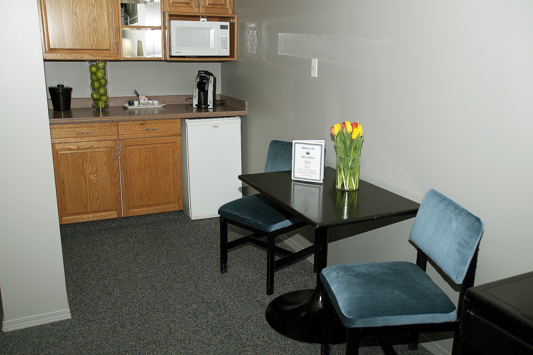 A small kitchenette with wooden cabinets, a microwave, and a coffee maker. There is a table with two blue chairs and a vase with flowers.