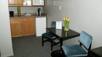 A small kitchenette with wooden cabinets, a microwave, and a coffee maker. There is a table with two blue chairs and a vase with flowers.