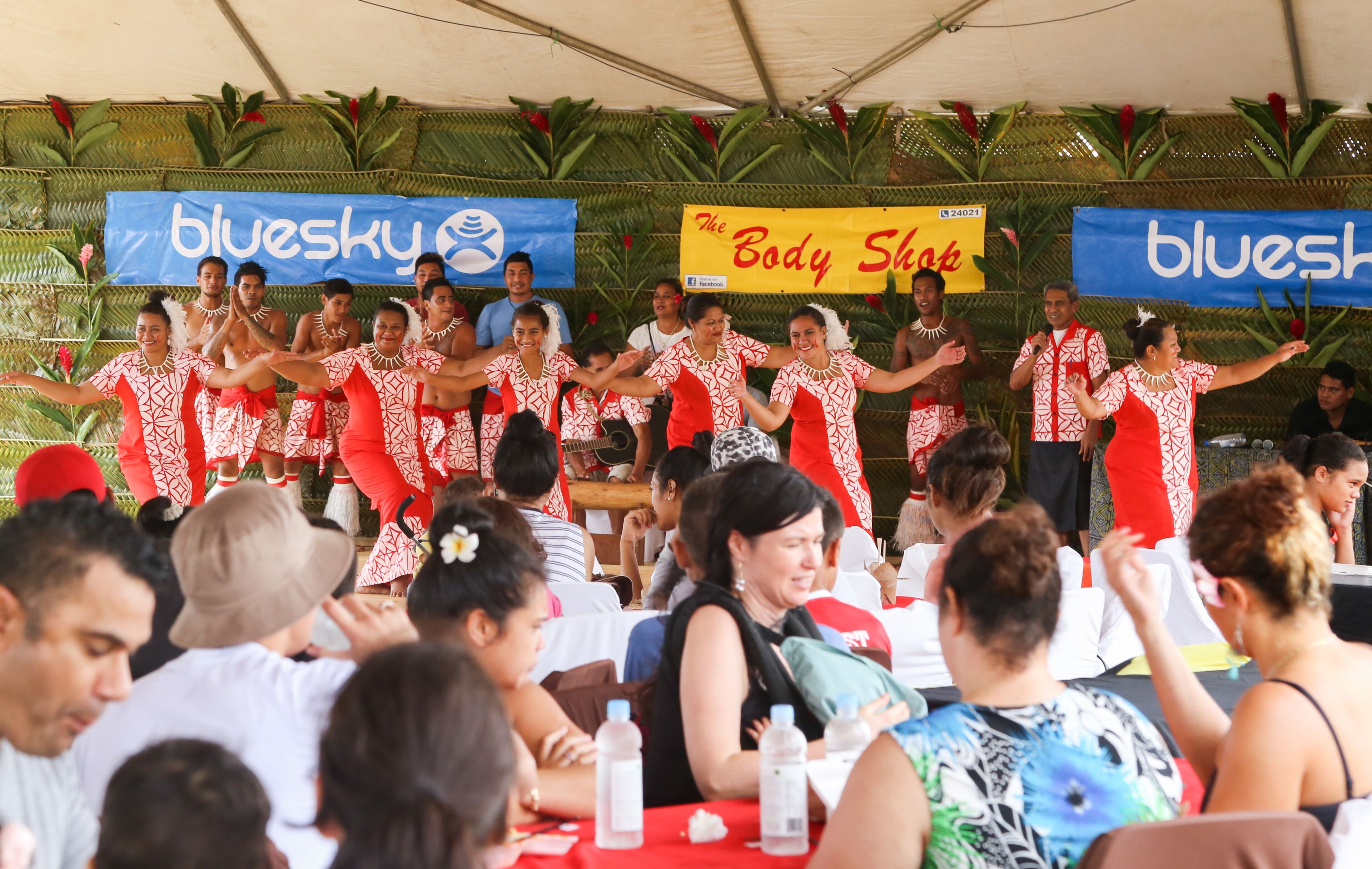 A group of people in red and white patterned outfits performing on a stage with a banner reading "The Body Shop."