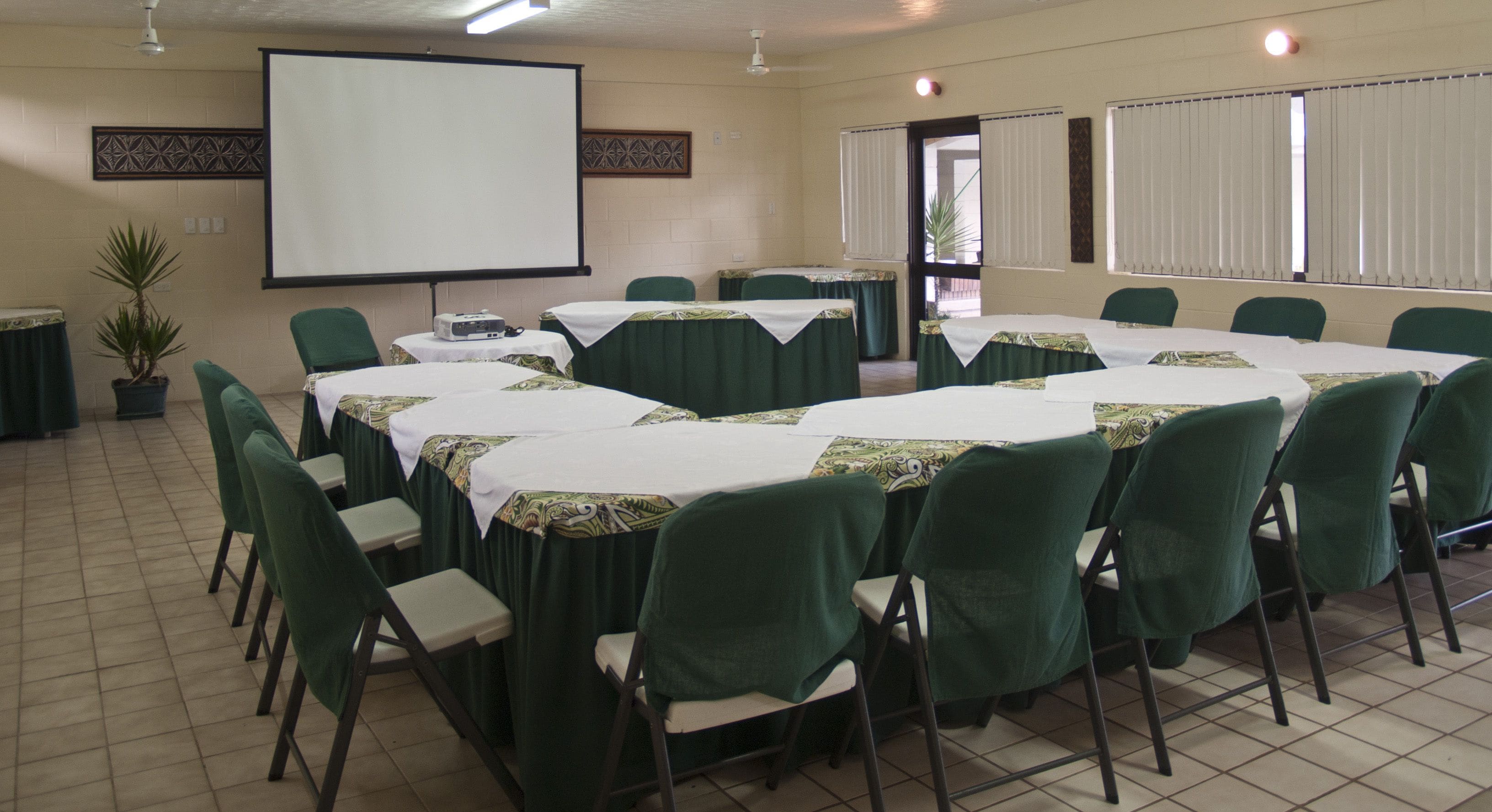 A conference room with a large screen, multiple tables with green-covered chairs, and a potted plant.