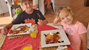 Two children seated at a table with breakfast plates, one with waffles and the other with pancakes, both topped with bacon and a dollop of butter.