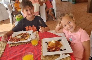 Two children seated at a table with breakfast plates, one with waffles and the other with pancakes, both topped with bacon and a dollop of butter.