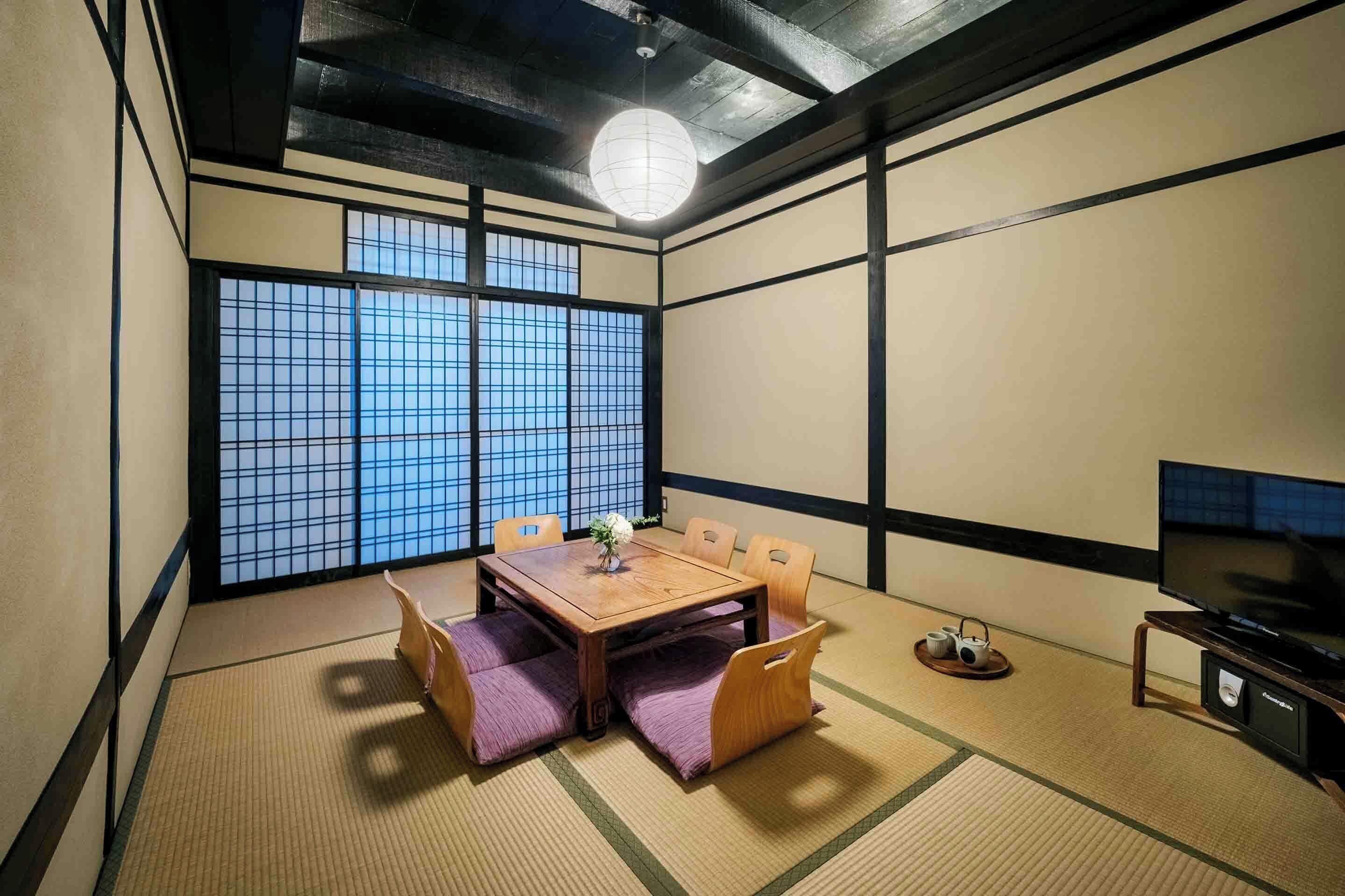 Ein traditioneller japanischer Raum mit Tatami-Böden, einem niedrigen Tisch mit Kissen, einem Fernseher und einem großen Fenster mit Gitterstruktur.