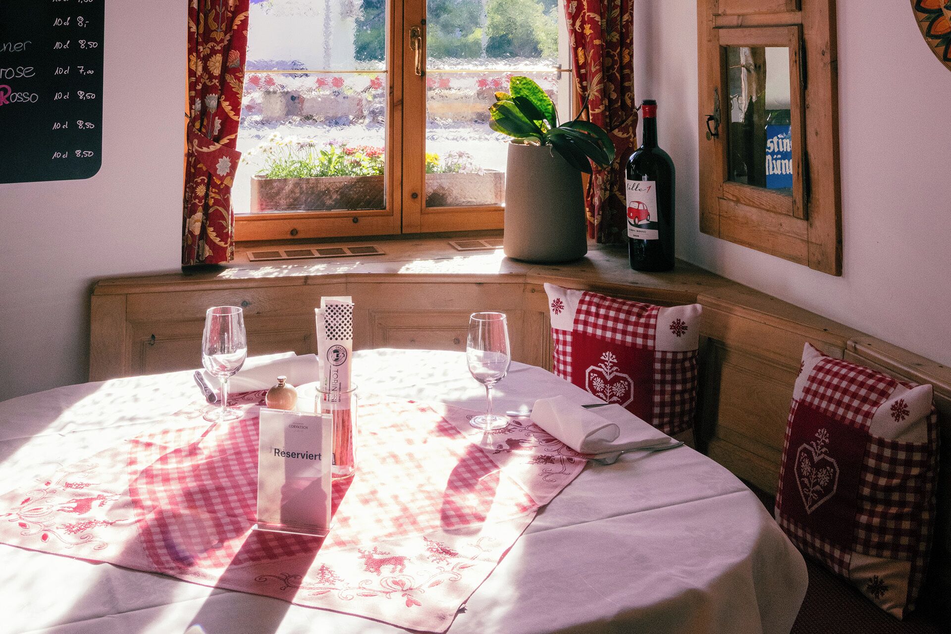 A table set for two with a checkered tablecloth, two wine glasses, a bottle of wine, and a menu on the wall.