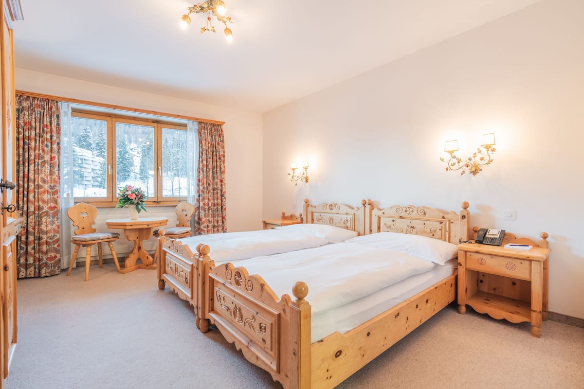 Une chambre à coucher avec deux lits en bois, une table de chevet en bois avec un téléphone, une chaise en bois, une petite table avec un vase de fleurs et une fenêtre avec des rideaux.