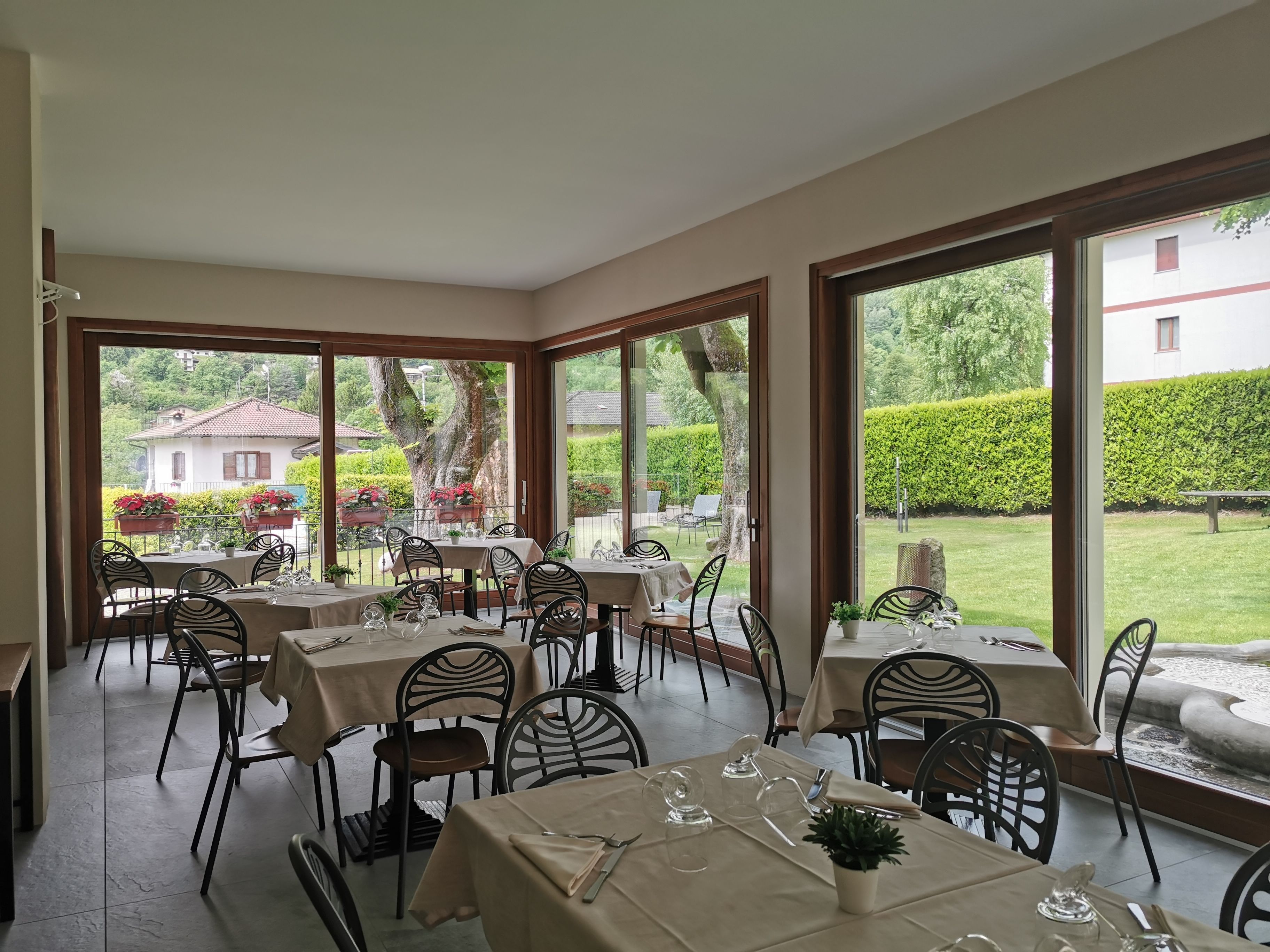 Un restaurant doté de nombreuses tables et chaises, de grandes fenêtres donnant sur un jardin et des bâtiments.