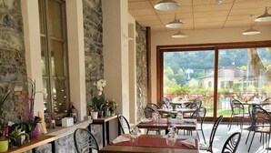 Un restaurante con paredes de piedra, ventanas amplias y mesas al aire libre. Las mesas están dispuestas con sillas y plantas en macetas.