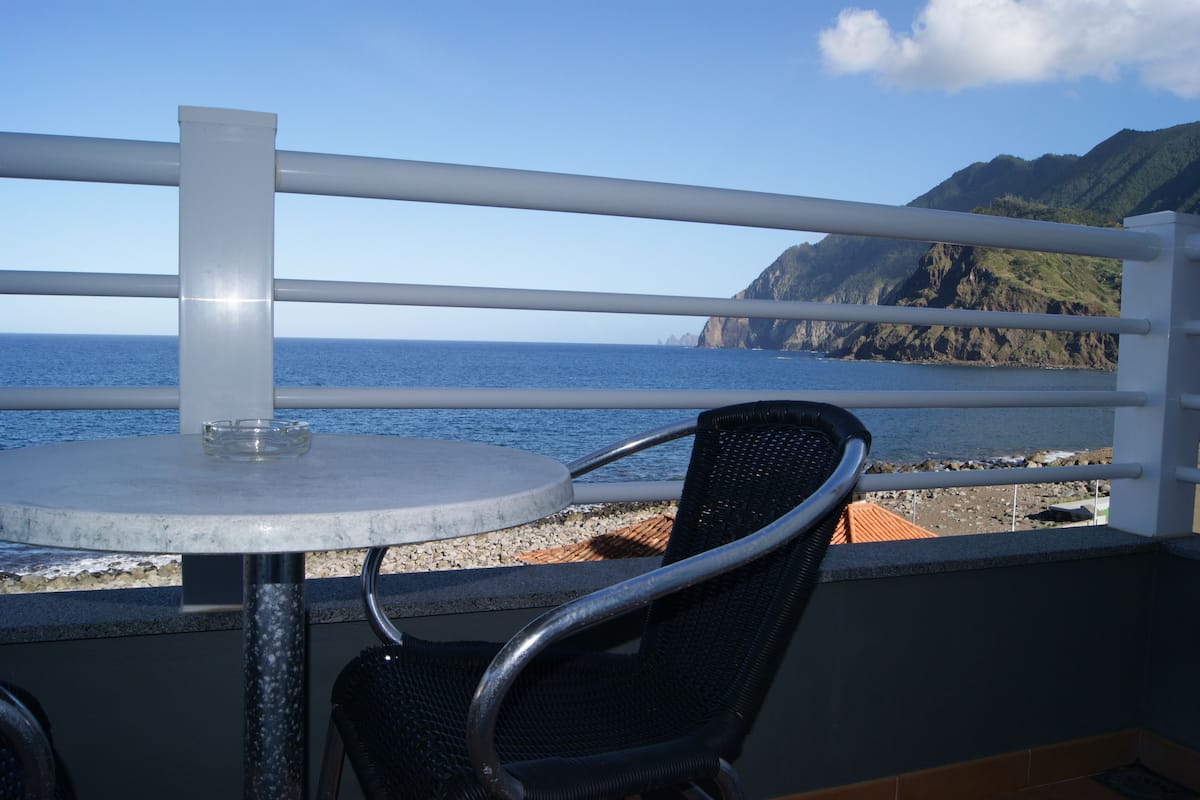 A balcony with a table and chair, overlooking a coastal view with rocky terrain and a clear sky.