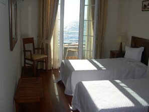 A room with two beds, a chair, a small table, and a view of the sea through large windows. - Hotel Costa Linda (Machico)