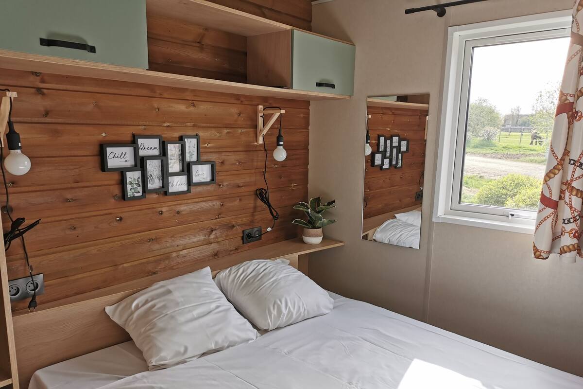 Quarto com cabeceira de madeira, cama com roupa de cama branca, mesa de cabeceira com abajur e janela com vista para o exterior.