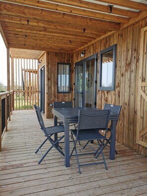 A covered wooden deck with a table and chairs, a sliding glass door, and a view of the outdoors. - Le Village de Cottages - Le Pôle du Cheval et de l'Âne (La Celle-Conde)