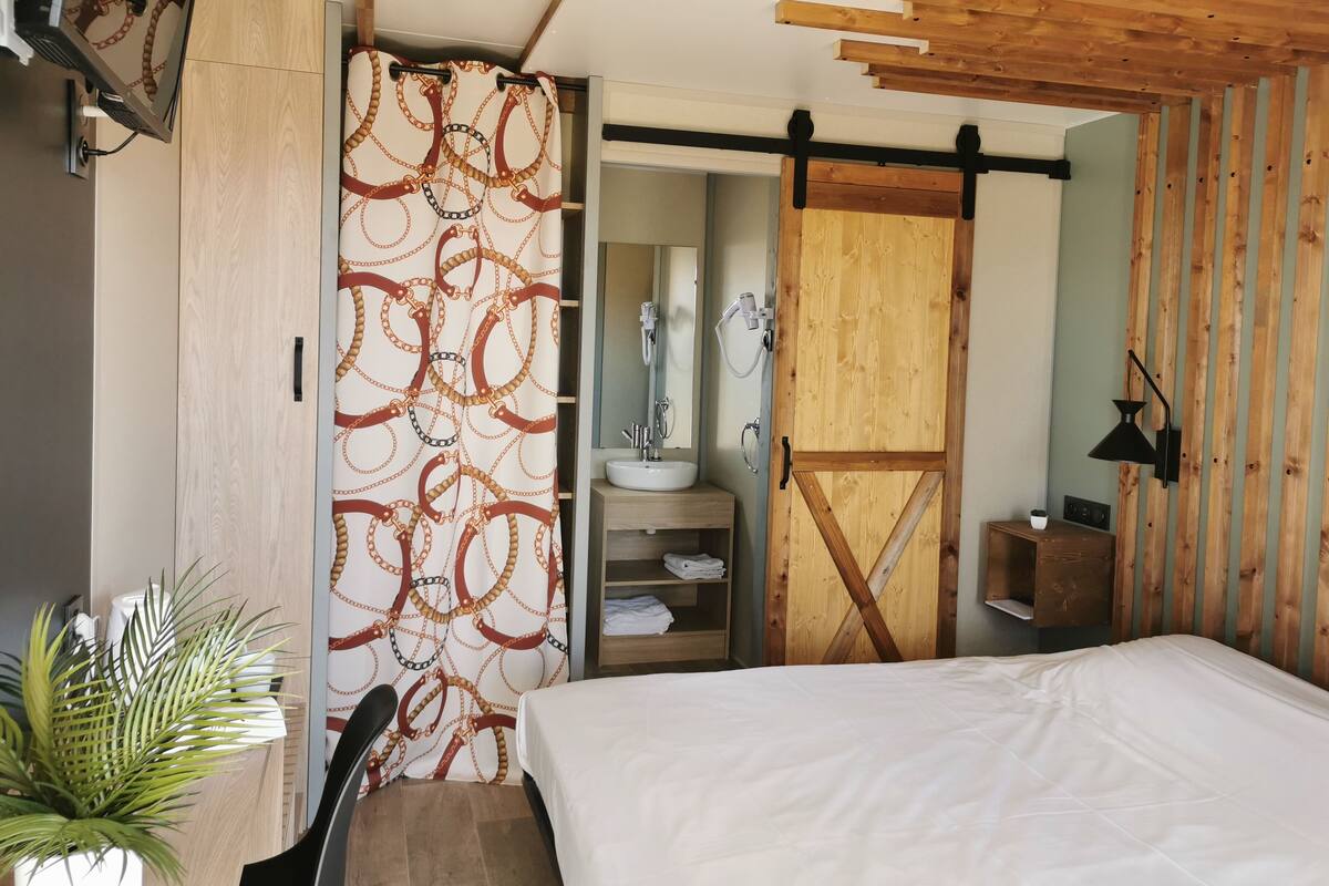 Quarto com cama, porta de madeira, banheiro com pia e espelho, e uma planta.