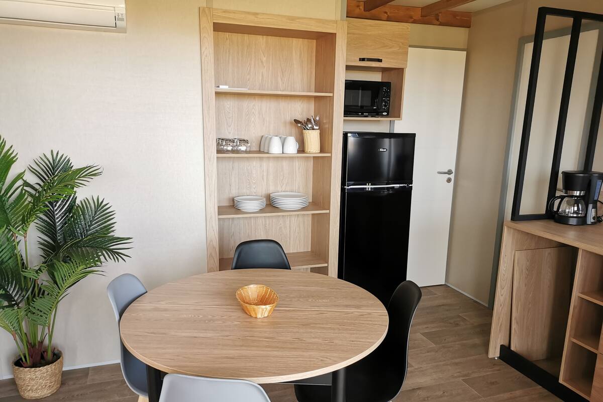 Cozinha compacta com geladeira preta, micro-ondas e cafeteira. Há uma mesa de jantar com cadeiras e um vaso de planta.
