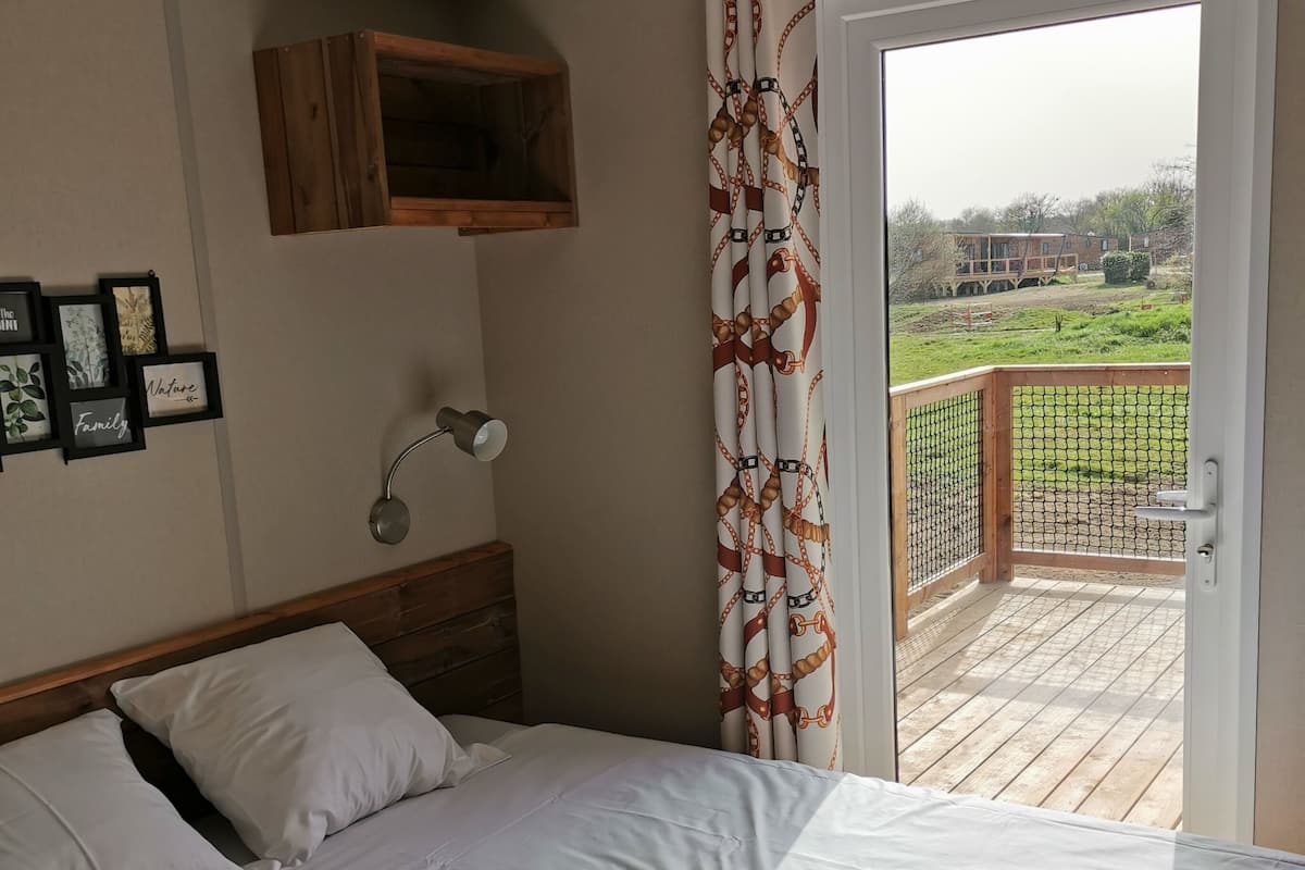 Quarto com cama, mesa de cabeceira, prateleira na parede e vista para uma varanda com deck de madeira e vegetação.