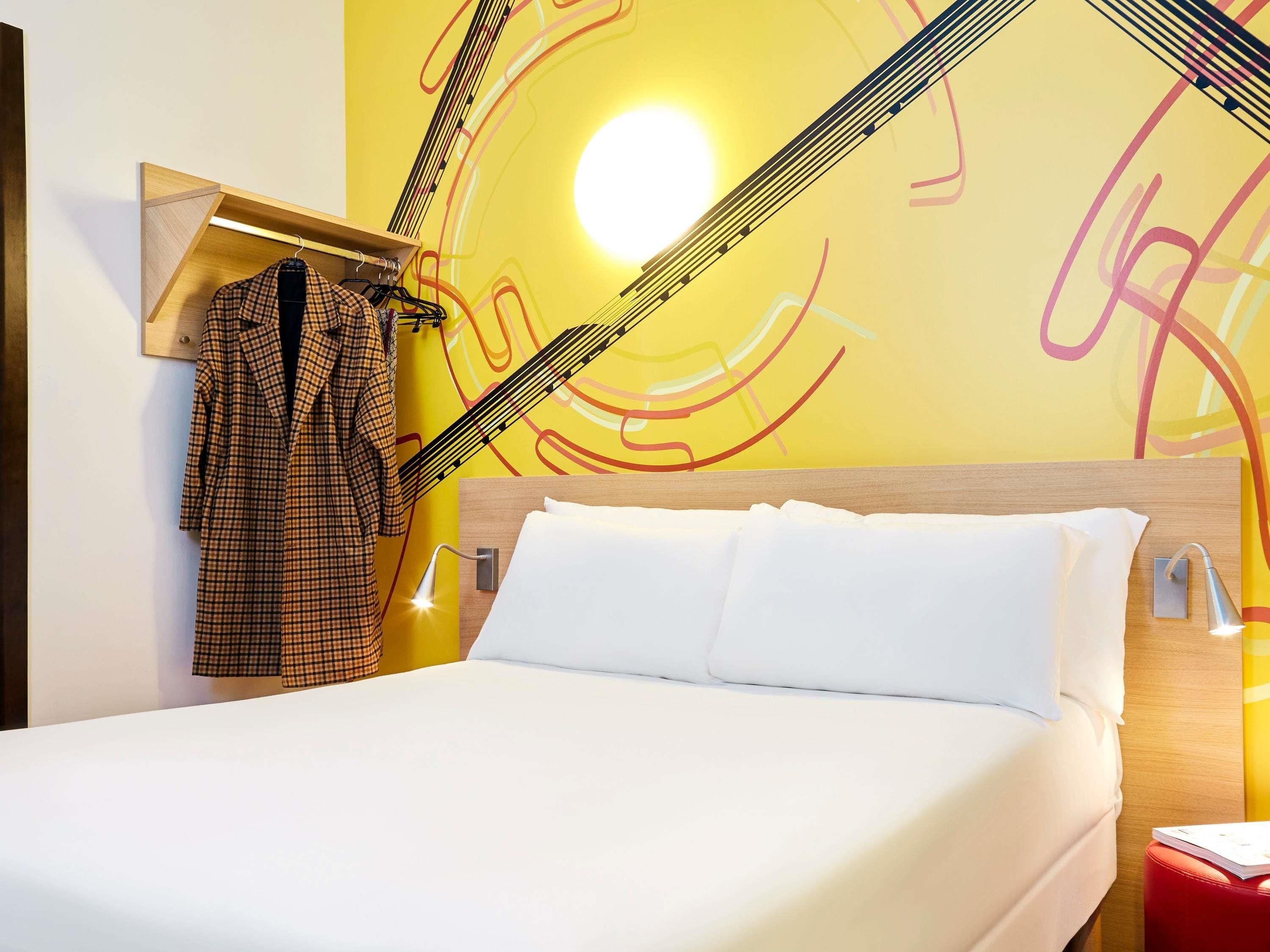 A hotel room with a bed, a hanging coat rack, a wall with abstract art, and a bright light source.
