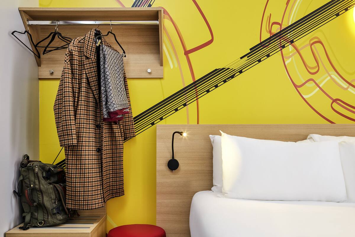 A room with a bed, a wooden headboard, a hanging coat rack, and a red stool.