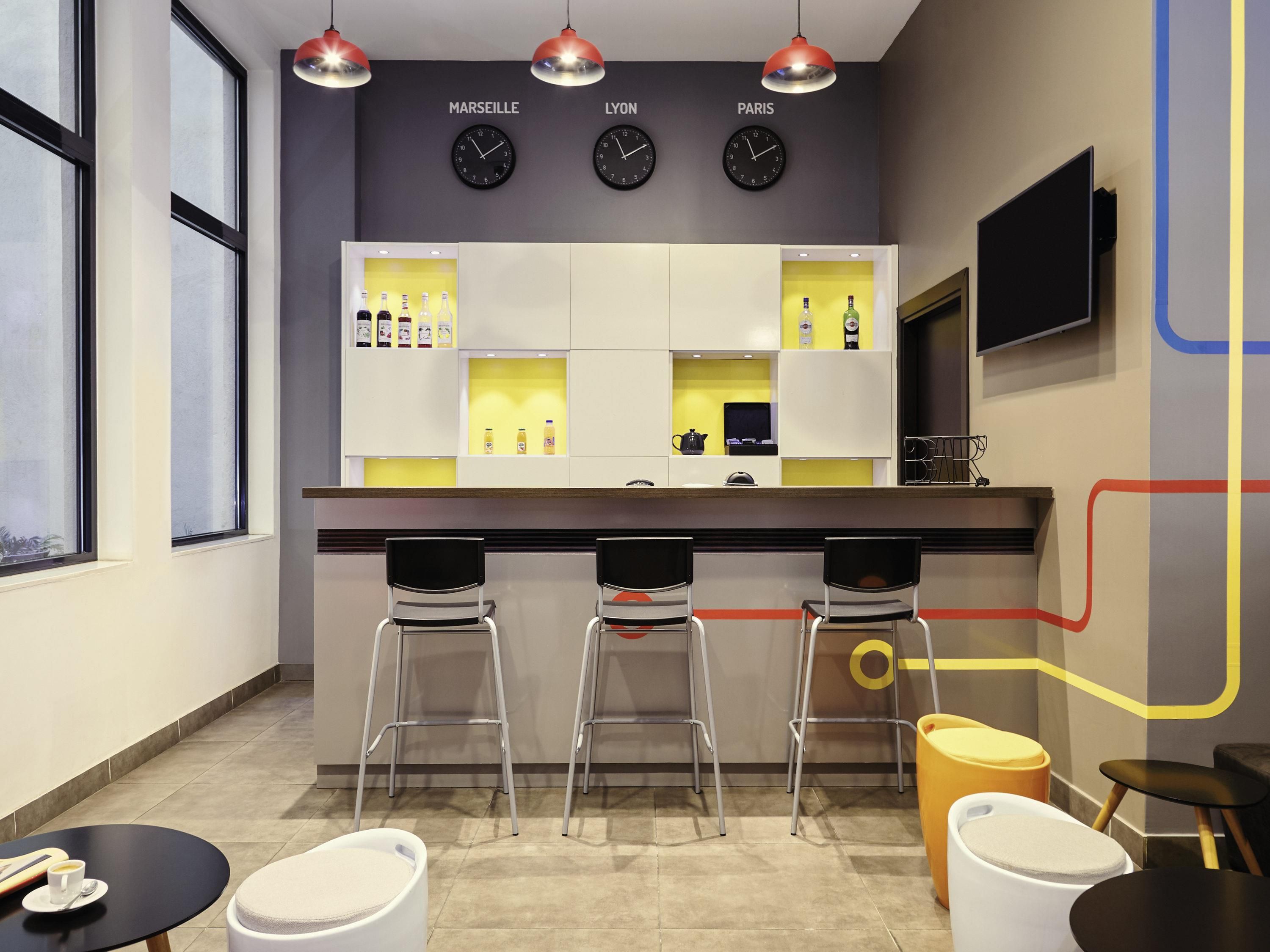 Un coin bar avec trois tabourets, un comptoir avec des bouteilles et des horloges portant les inscriptions « MARSEILLE », « LYON » et « PARIS ».