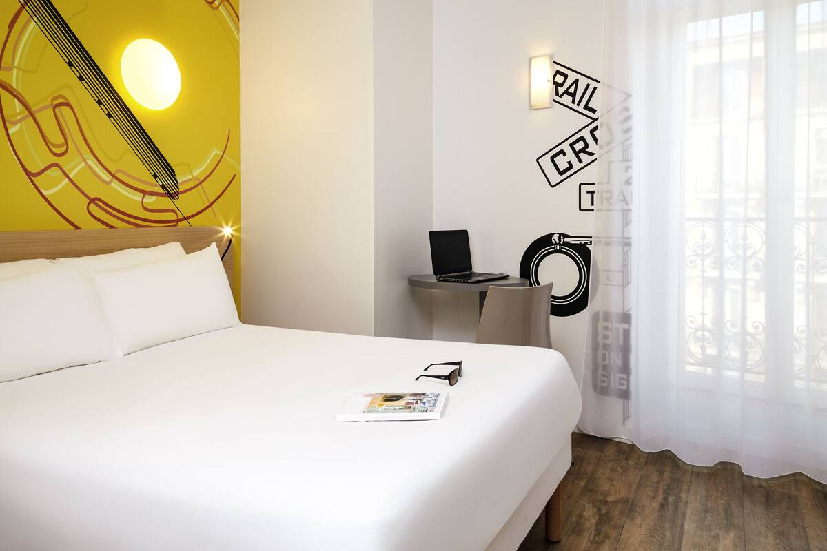 A hotel room with a bed, a small desk with a laptop, a wall clock, and a window with sheer curtains.