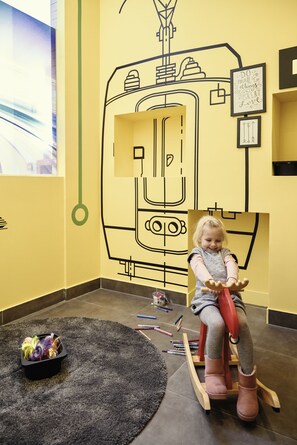 Un enfant est assis sur une chaise à bascule dans une pièce aux murs jaunes, ornée d’une grande fresque abstraite.