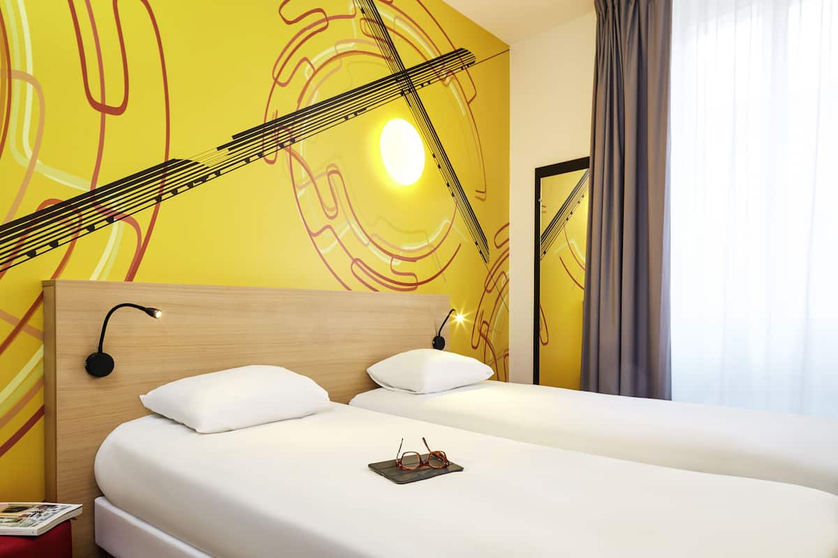 A hotel room with a bed, two pillows, a pair of glasses on the bed, a nightstand with a lamp, and a wall with abstract art.