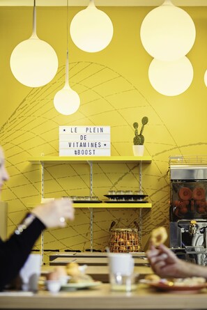 L’intérieur d’un café avec un mur jaune, des suspensions lumineuses, une enseigne indiquant « LE PLEIN DE VITAMINES #BOOST », une machine à café et un panier de pain.