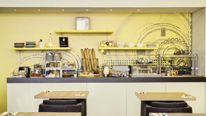 Um café moderno com esquema de cores amarelo e branco, apresentando um balcão com diversos itens alimentares e um relógio na parede. Há duas mesas com cadeiras em frente.