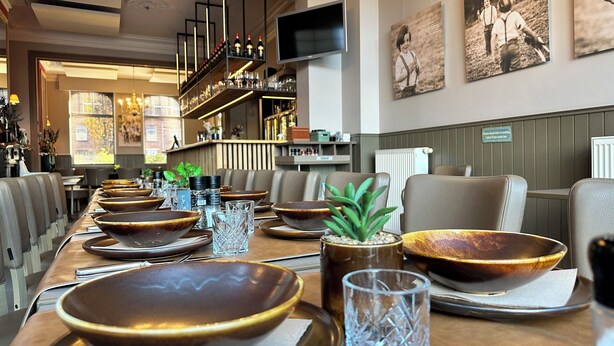 A restaurant with a long dining table set with bowls, glasses, and a potted plant. There are chairs around the table, and a bar with shelves of bottles in the background. - 't Oud gemeentehuis Poelkapelle (Langemark-Poelkapelle)