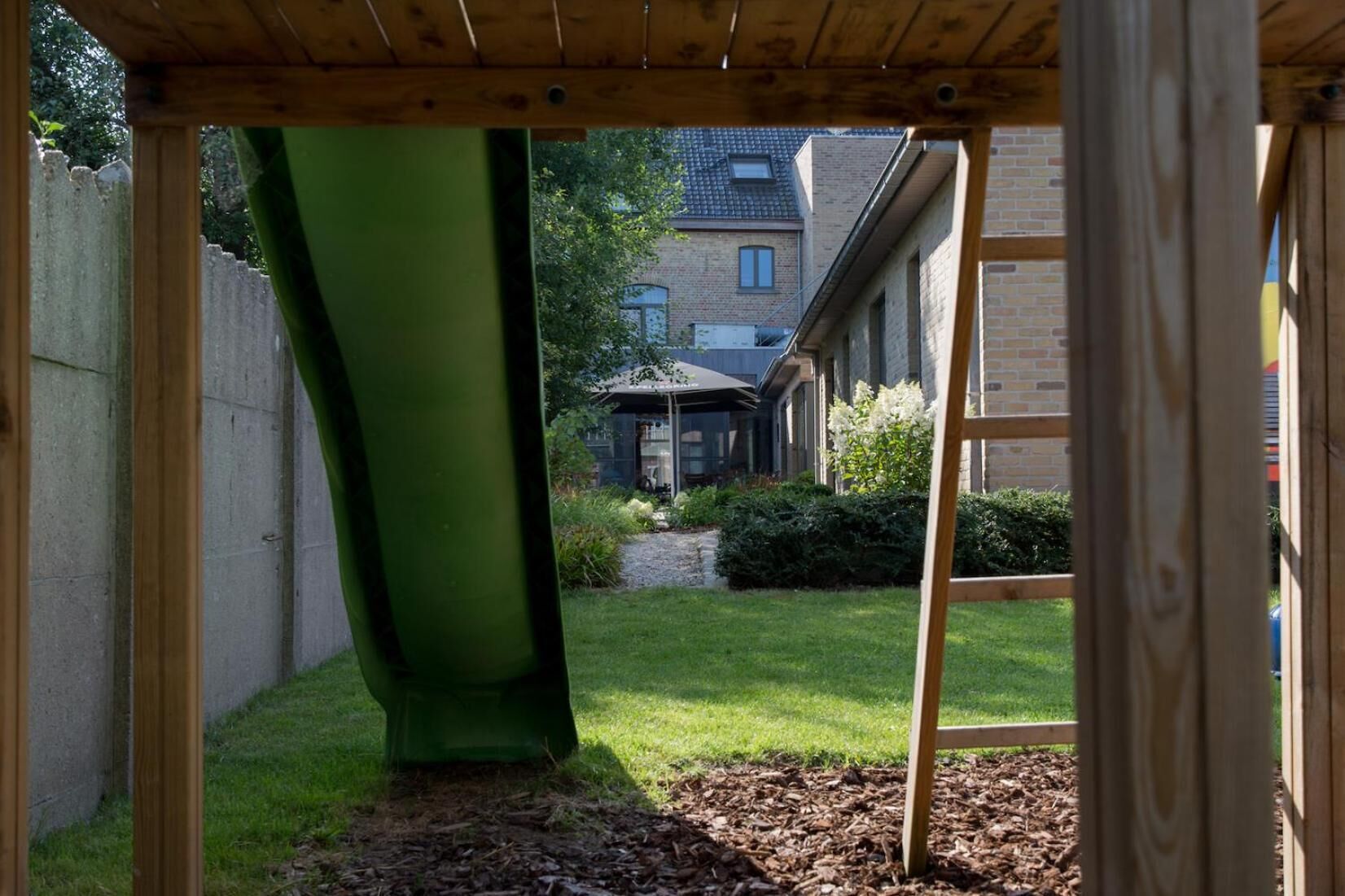 A wooden structure with a green slide and a ladder, leading to a backyard with a lawn and a brick building in the background.