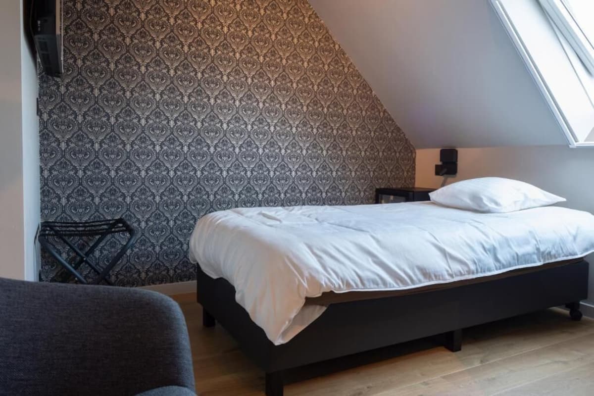 Une chambre à coucher avec un papier peint à motifs, un lit recouvert de draps blancs, un plancher en bois et une lucarne.