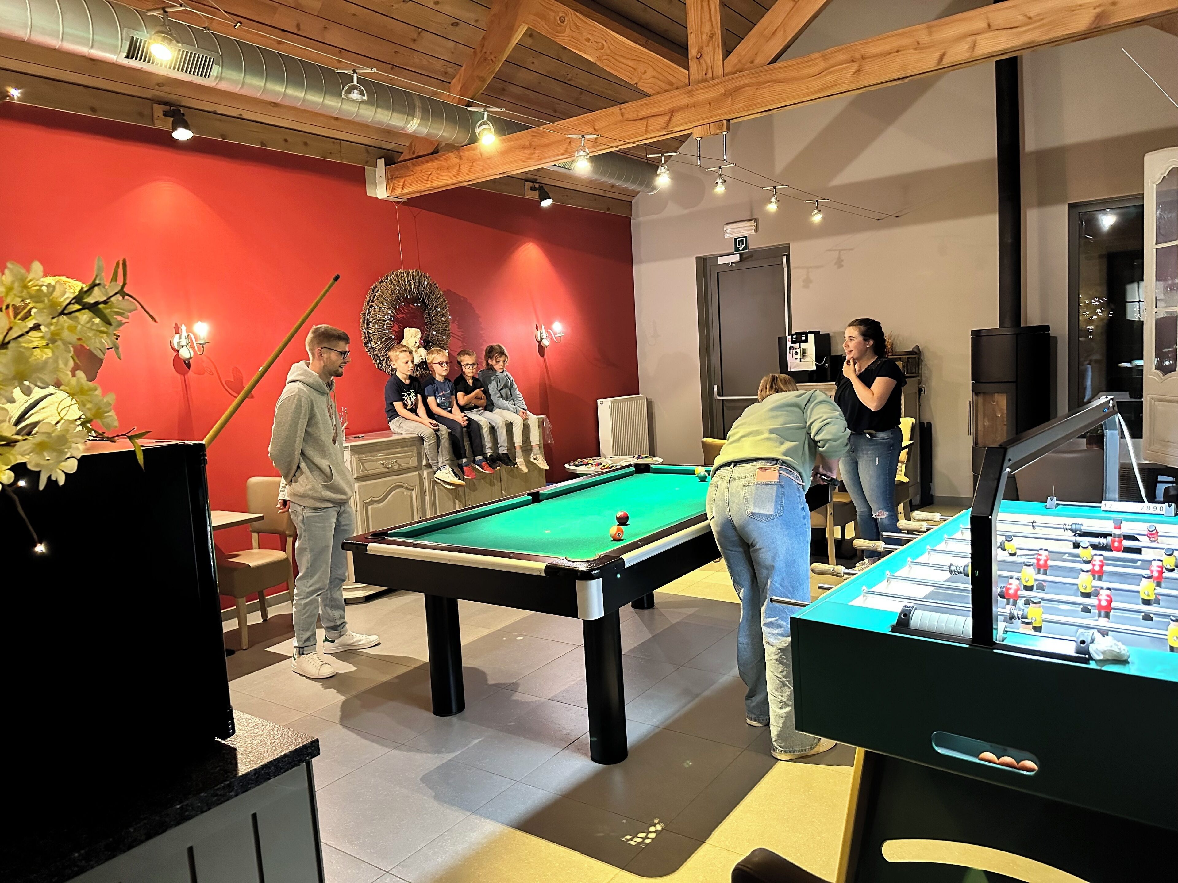 A billiards room with a pool table, a foosball table, and a red wall with a wreath decoration.