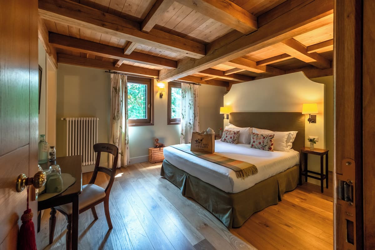 Un dormitorio con techo de madera, una cama con cabecera, una mesita de noche, una silla y una ventana con cortinas.